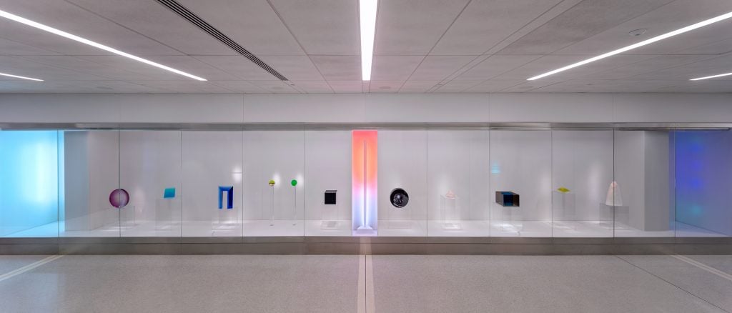 The image shows a contemporary art installation in an airport. The display features a variety of modern art pieces, including geometric shapes, colored lights, and abstract forms, all enclosed in a glass case. The background is minimalistic, with clean lines and soft lighting that highlights each artwork, creating a serene and contemplative atmosphere.