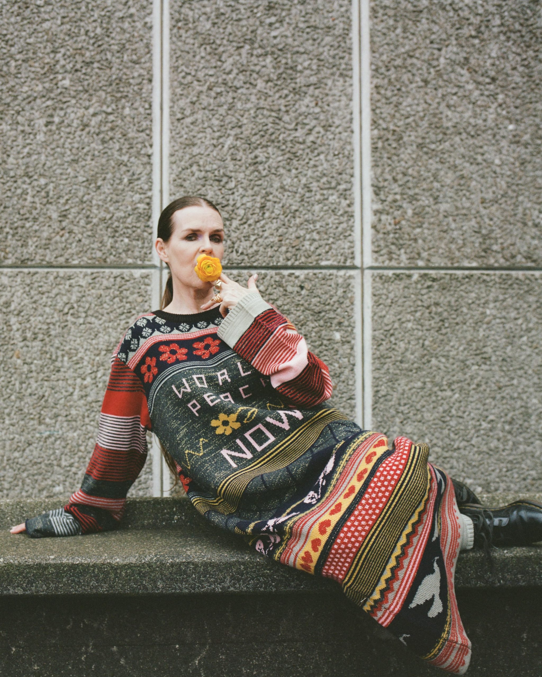 the artist Linder wears a knee length colorful sweater reclines on a marble bench with a floor in her mouth