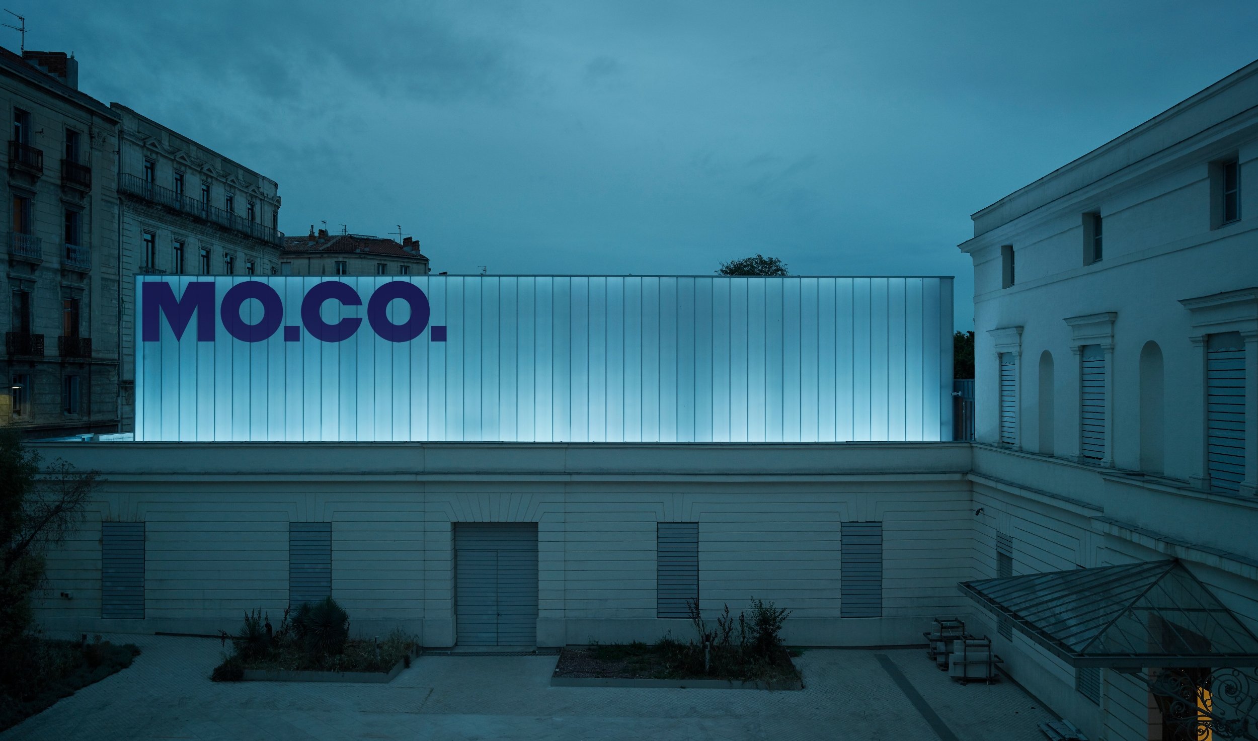 a pale museum building with the letters "MO.CO" written in big blue capital letters, it is quite dark and gloomy, probably evening and cloudy skies