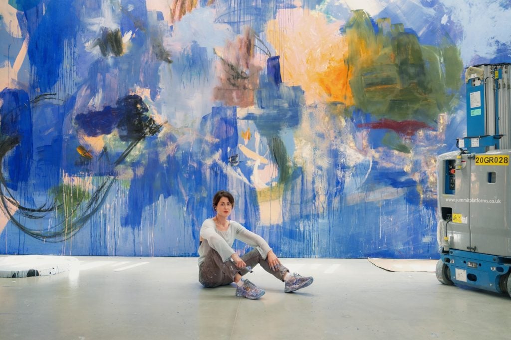 a woman sits in front of a large painted abstract mural with predominant colors of blue