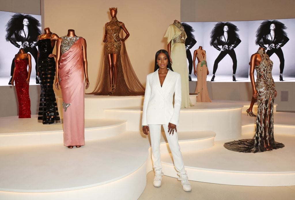 a woman stands in front of a group of mannequins wearing various gowns, the woman is wearing a white suit and there are some images of her model that are projected onto the background