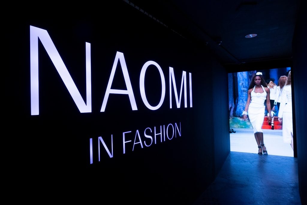 a long dark corridor with the text "naomi in fashion" written in white along it and at the end a projection of a woman strutting on a catwalk