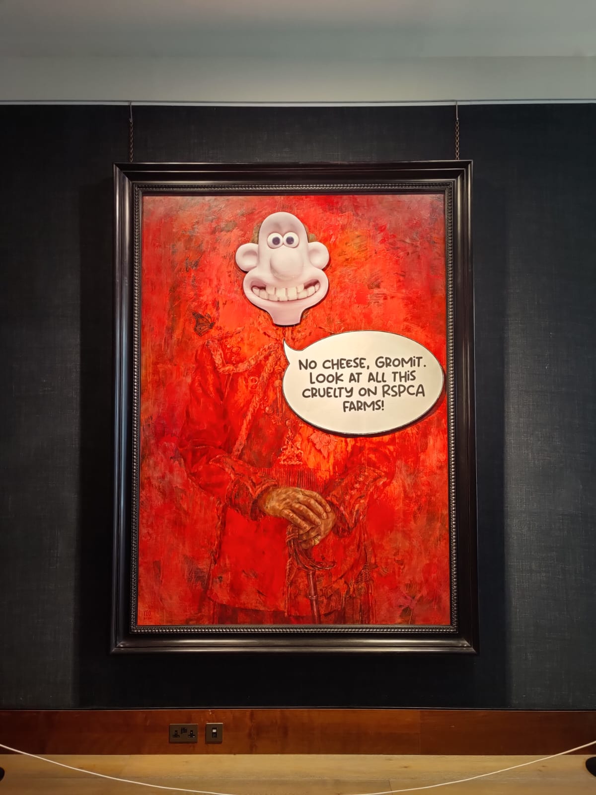 A framed artwork on a dark wall features an abstract, predominantly red painting with a three-dimensional, comically exaggerated head of Wallace from "Wallace and Gromit" protruding from the top. Below the head is a speech bubble that reads, "No cheese, Gromit. Look at all this cruelty on RSPCA farms!"