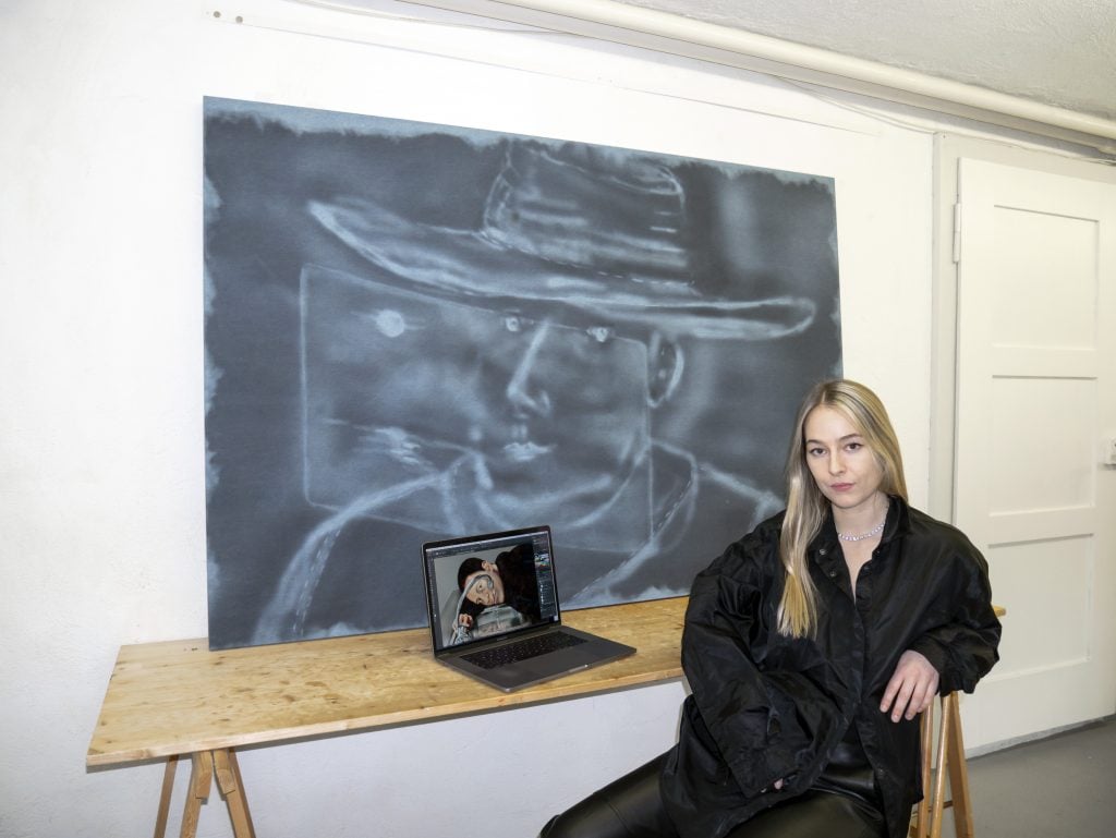 a woman with blonde hair sits at a wooden table. she is a wearing a bit black buttondown shirt and black paints. a laptop is on the table and a chalk-like drawing of a man in a cowboy hat rests against a wall