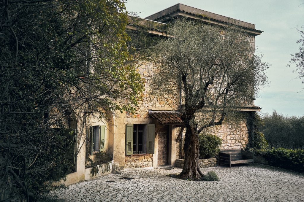 The exterior of Picasso's final home in Mougins, located in the French Riviera. The rustic stone house features green shutters, a cobblestone courtyard, and is surrounded by lush foliage and an olive tree, reflecting the serene and picturesque setting where Picasso spent the last years of his life.