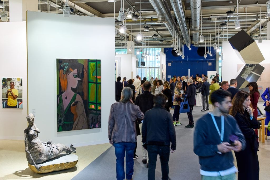 A group of people walk through an art fair. there is a painting and a sculpture to the left of them