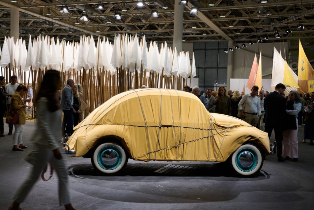 a VW beetle wrapped in a mustard yellow cloth stands in front of a stand of white flags