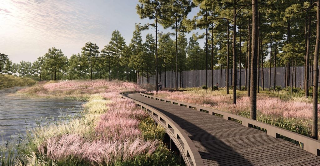 A rendering of the "Passage of Time Pavilion" view from the pond at Longleaf Art Park. The image shows a boardwalk winding through a landscape of pink and green grasses by the water's edge, surrounded by tall pine trees. In the background, a modern pavilion structure is visible among the trees, blending into the natural environment. The scene is tranquil and scenic, emphasizing the integration of architecture with nature.