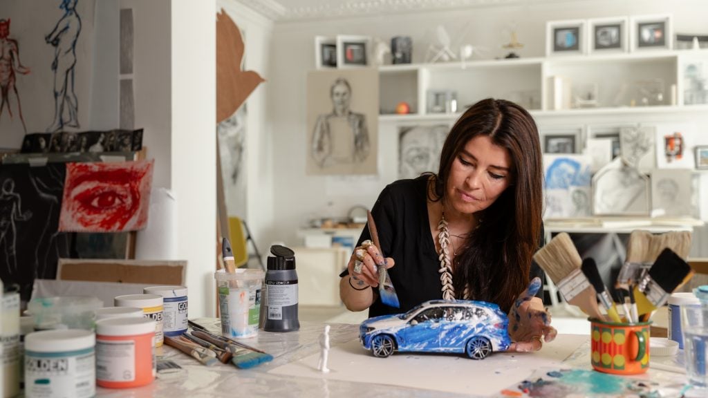 a woman inhabits a large studio full of art all over the walls and pots of paint etc, she is seated at a desk and is painting a small model car the colour blue