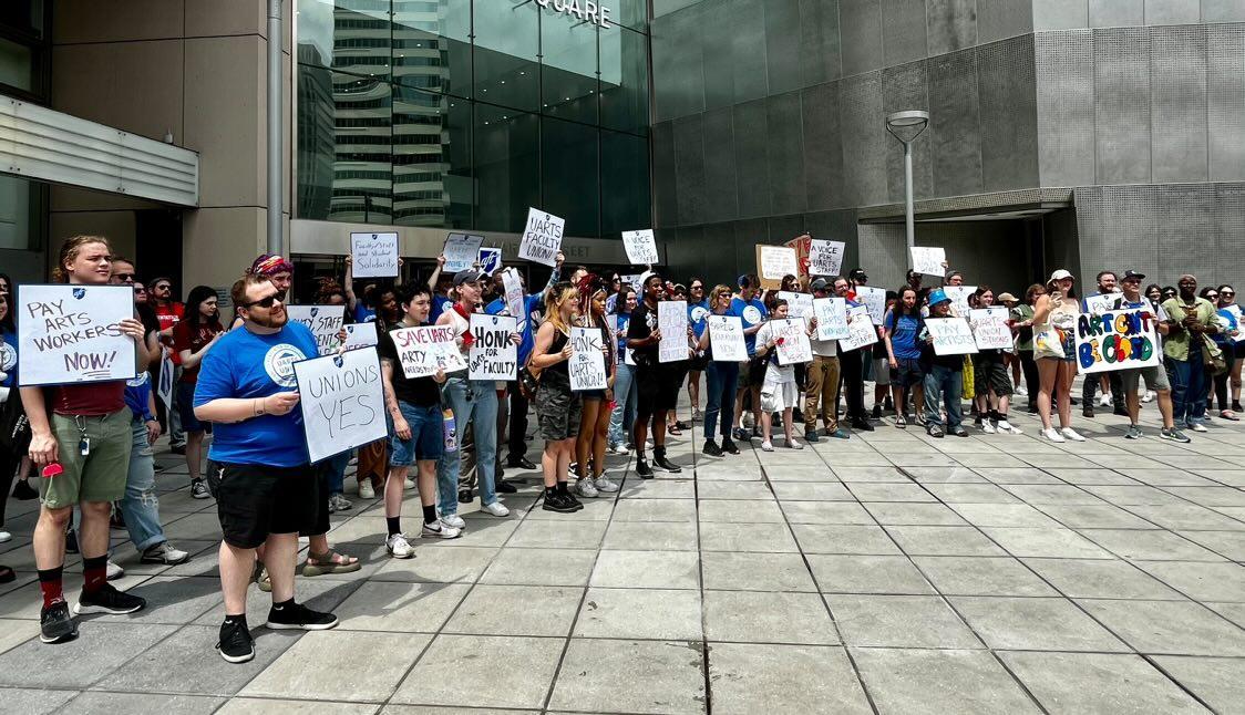 A group of people gathered outside a modern building, holding various signs advocating for arts workers' rights and union support. The signs include messages like "Pay Arts Workers Now!", "Unions Yes", and "Honk for UArts Faculty Union." The demonstrators appear engaged and united in their cause.