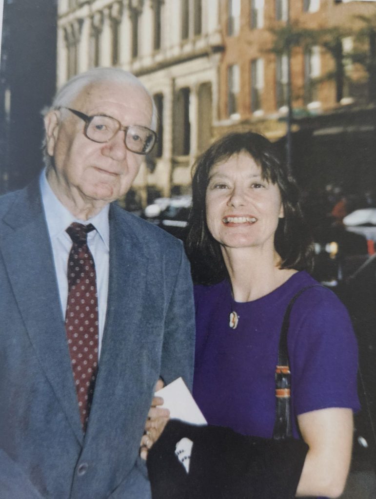 An elderly man with glasses, wearing a gray suit and red tie, stands next to a smiling woman with shoulder-length dark hair in a purple dress. The photo appears to be taken outdoors on a city street in front of buildings