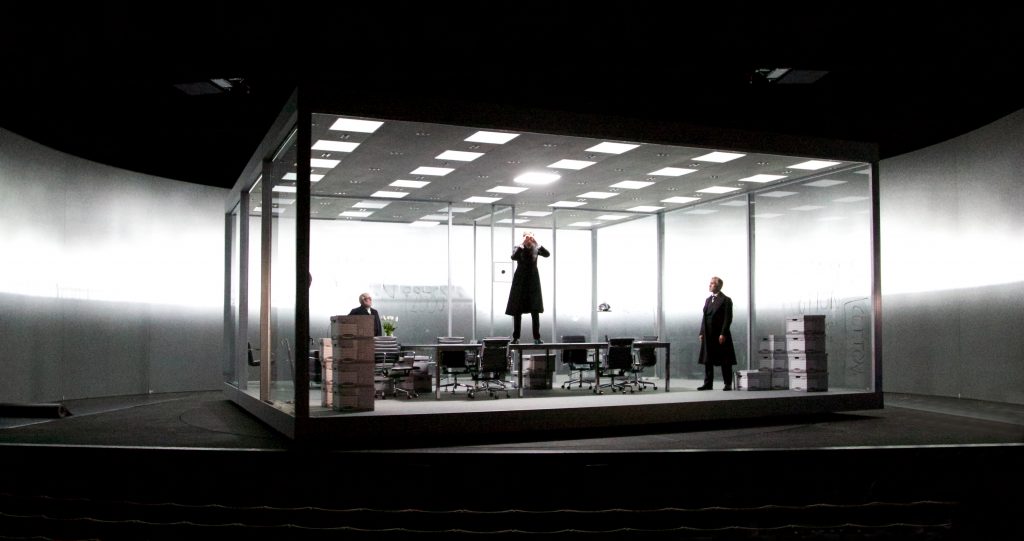 on a dark theatre stage we see a large glass box that is brightly lit in the manner of and office. it is in habited by a few suited figures and has chairs and desks. a faint new york style skyline can be seen behind