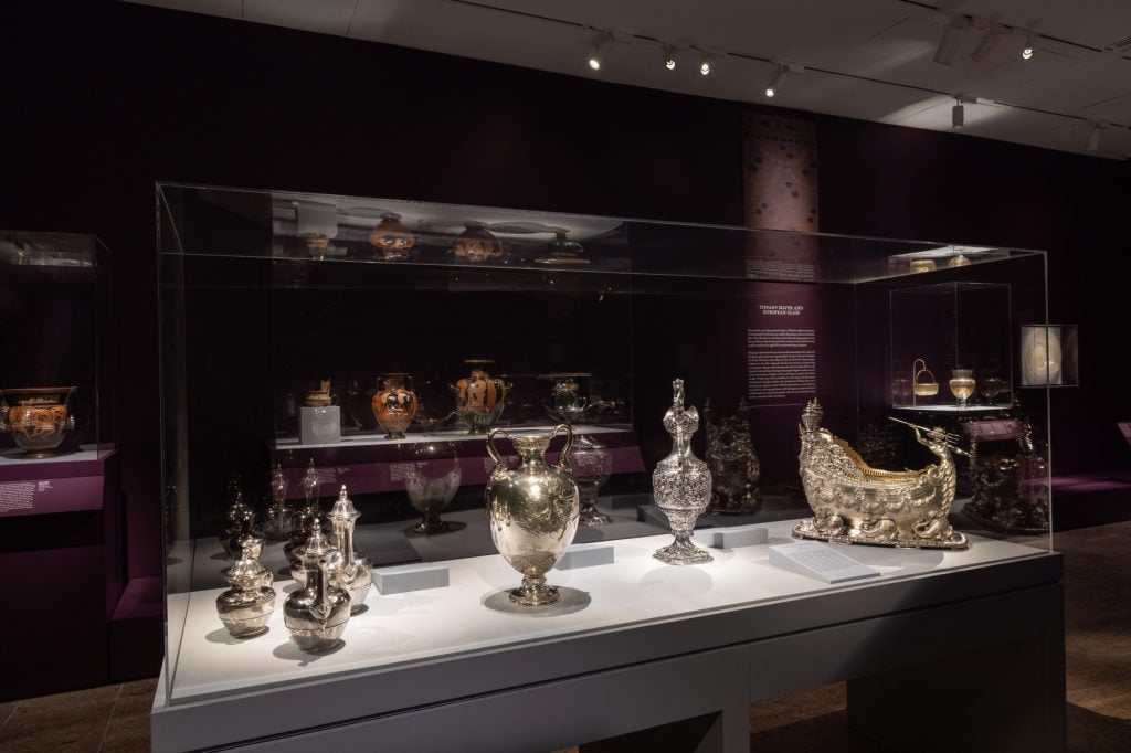 a dark gallery space with exquisite items of silverware in spotlit glass cases