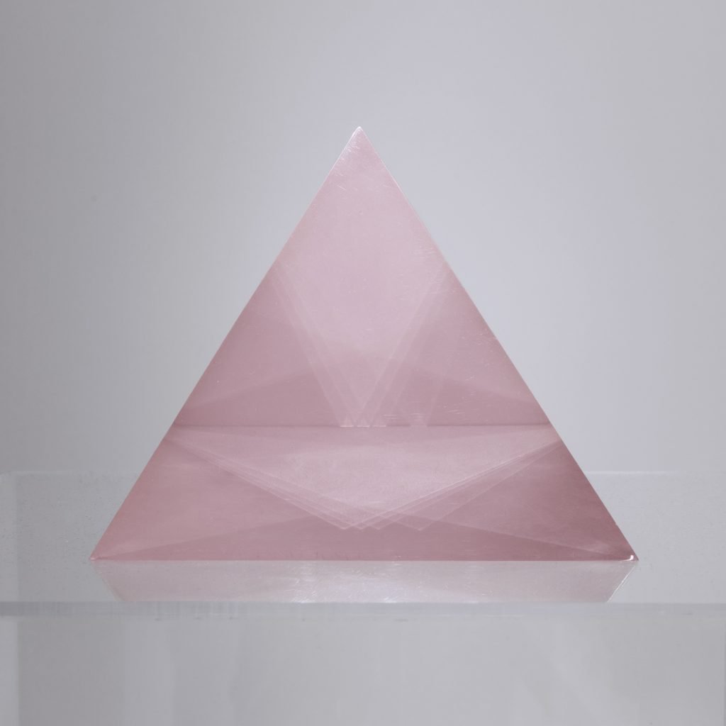 The image shows a triangular prism made of a translucent pink material. The prism has a smooth surface and reflects light subtly, creating a soft, delicate appearance. The object is displayed on a clear pedestal against a plain background, emphasizing its geometric simplicity and aesthetic elegance.