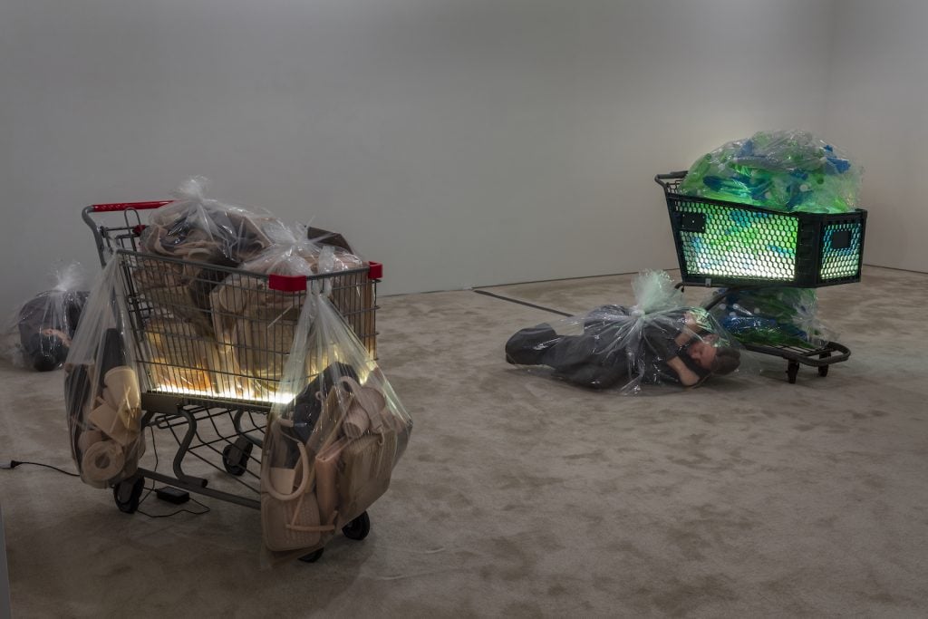 Two shopping carts hold glowing contents in a gray-walled gallery. A man on the floor appears to be wrapped in a plastic bag.