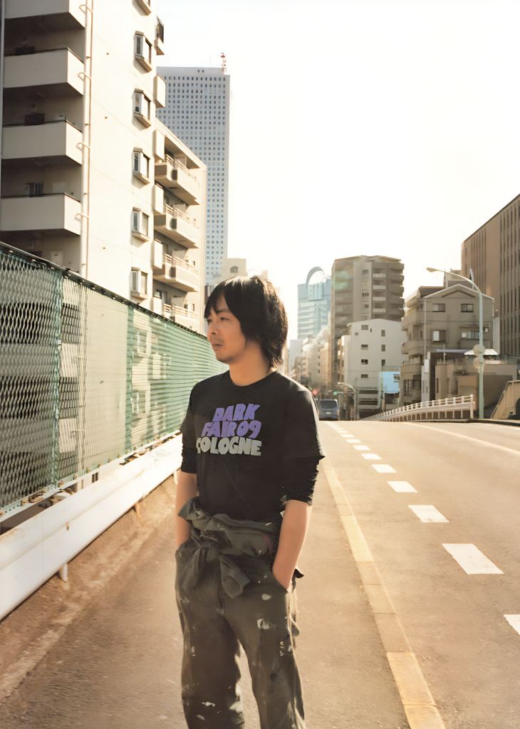 An East Asian man standing on the road surrounded by high risings.