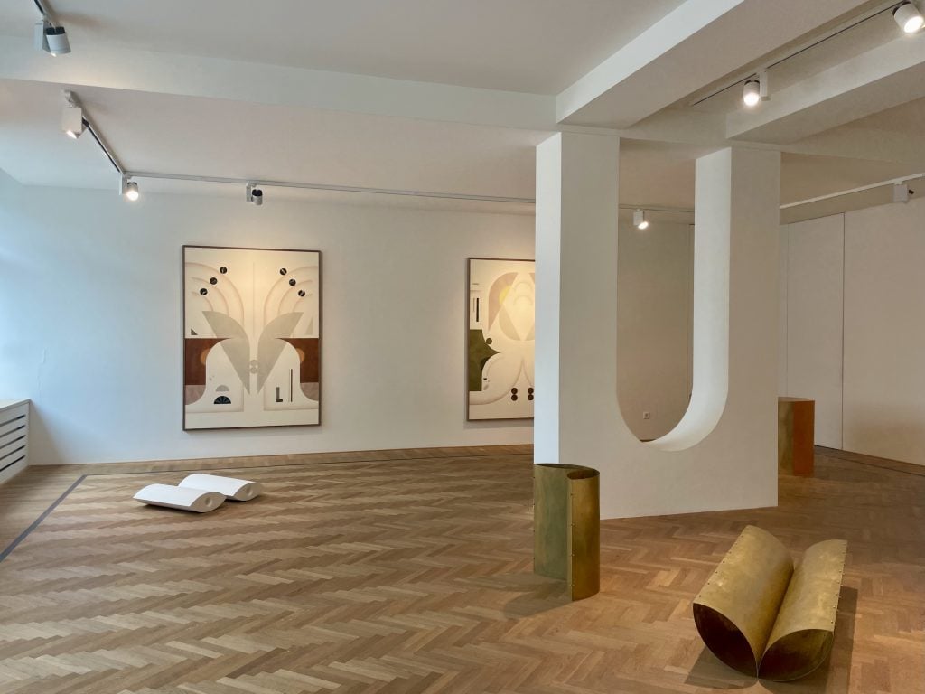 a photo of a gallery with white walls and a wood floor. two abstract works are on the far wall, on the floor there are several medium sized abstract sculptures