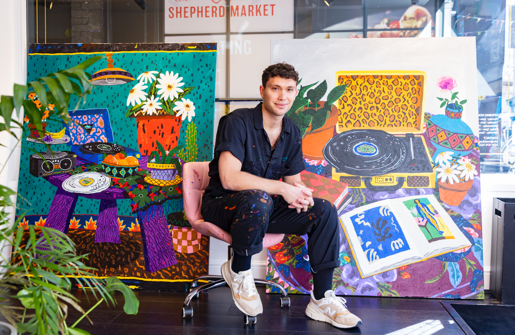 Portrait of the artist Cooper siting in front of a series of his paintings in a salmon colored chair with castors wearing white and pink steakers, a short sleeve navy button down, and black joggers spattered with paint.