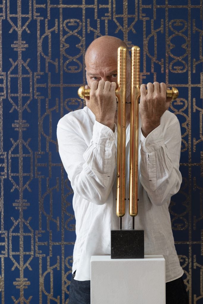 Portrait of the artist Kendell Geers peering out from behind a gold sculpture that he has his hands gripping, in front of a blue background with a pale metallic geometric pattern overlaid.