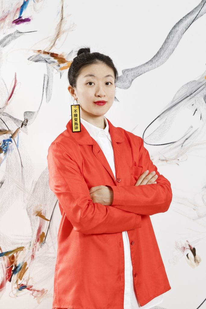 Portrait photo of artist Xiyao Wang standing in front of one of her abstract paintings wearing a bright coral blazer and red lipstick with hair in a bun.