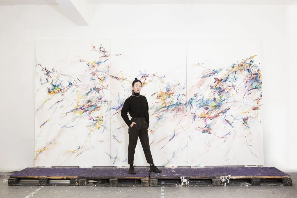 Portrait photo of the artist Xiyao Wang wearing all black standing in front of a large-scale mostly white abstract painting resting on wooden pallets.