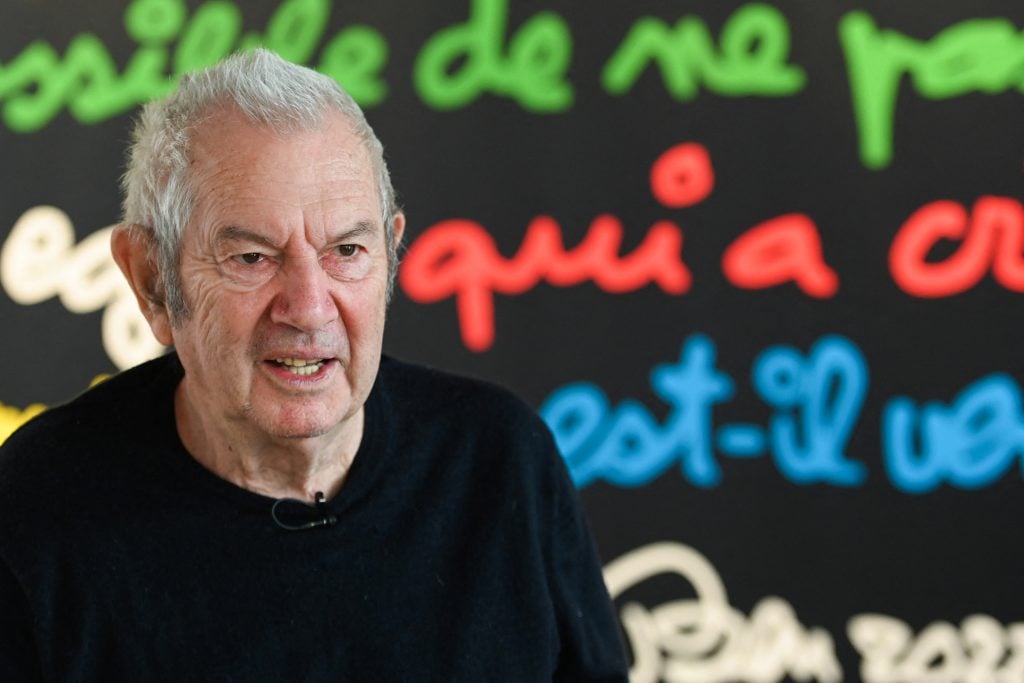 an aging man is seated in front of a background of colourful handwriting in French