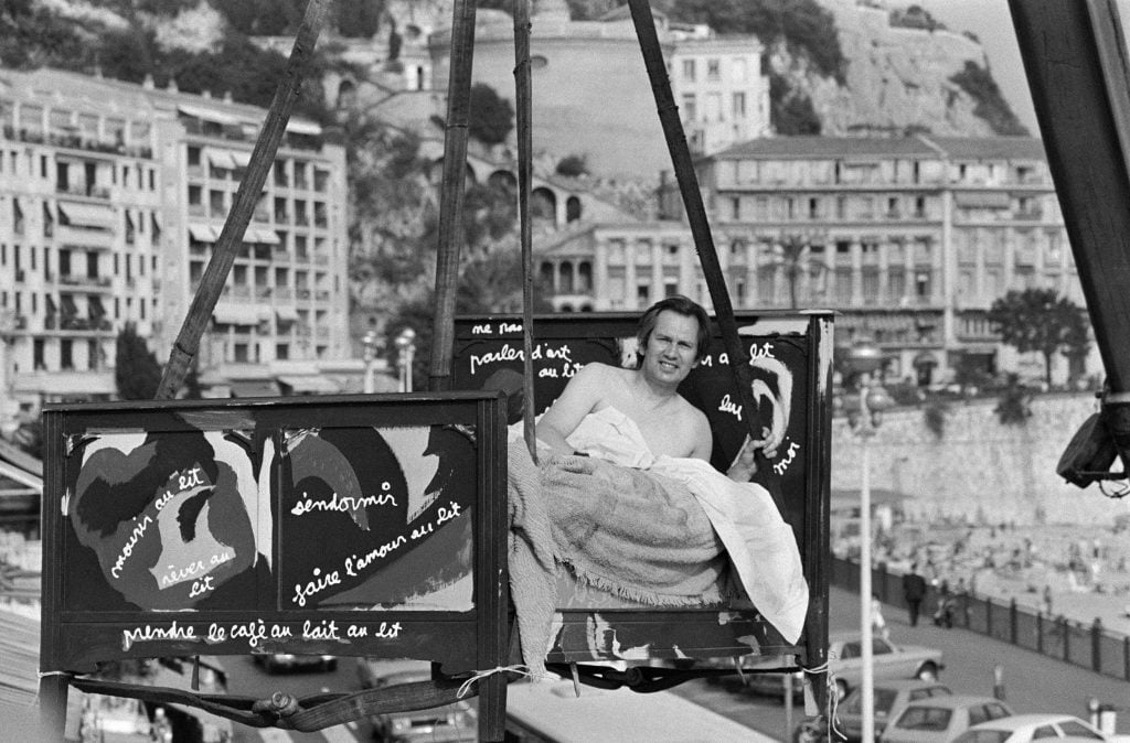 a black and white archival photo of a man in a wooden bed with slogans written on it in paint that is being suspended over the city of Nice