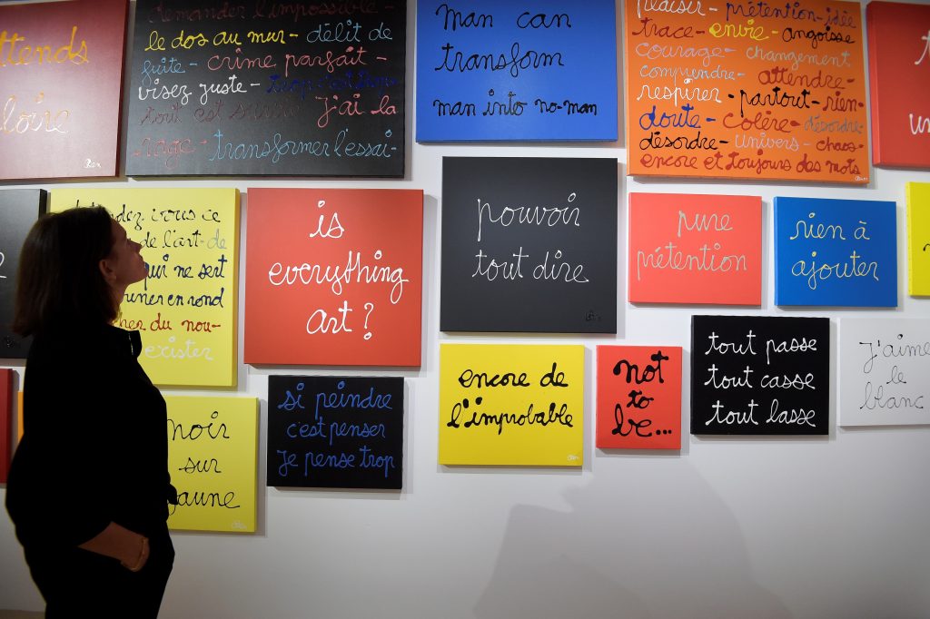 a lot of colorful pictures on the wall with white writing on them, some of the slogans are in english but most are in French, a woman stands to the side looking up at them 