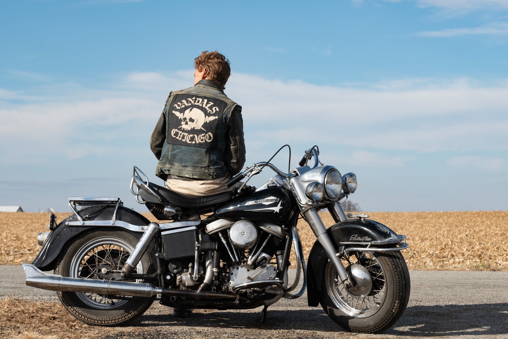 A still from the film The Bikeriders showing the back view of a man, clad in a leather jacket with the words "Vandals Chicago," sitting on a motorcycle