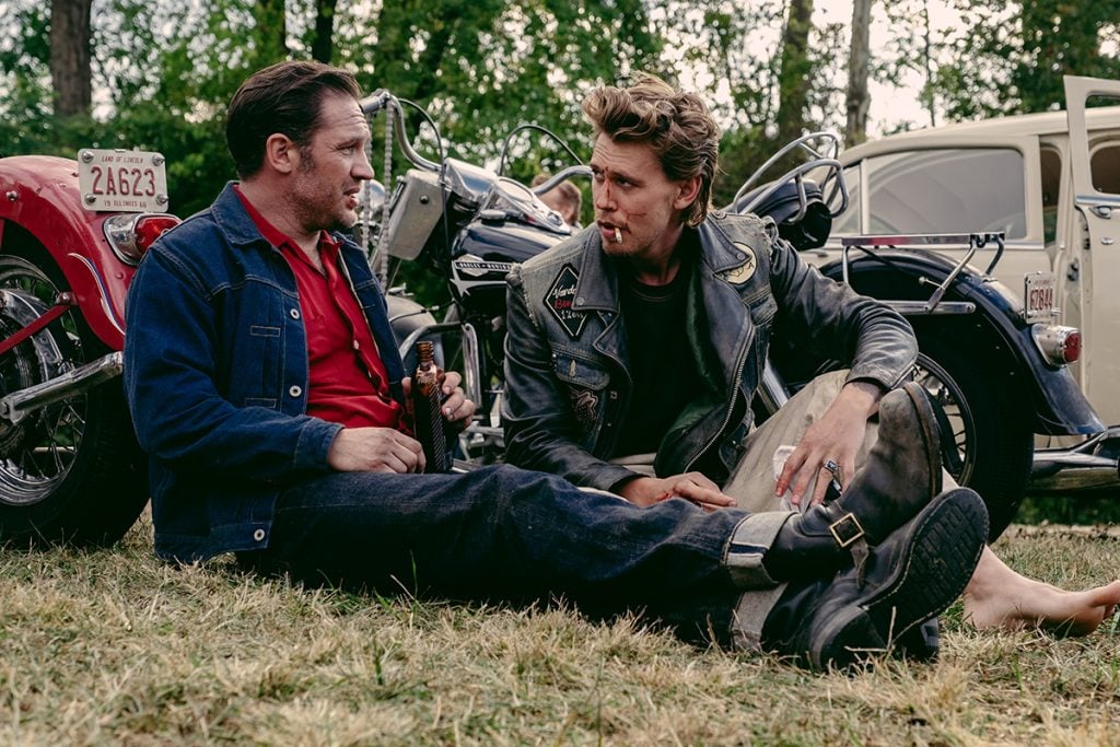 A still from the film The Bikeriders showing two men sitting on a grassy patch beside motorcycles