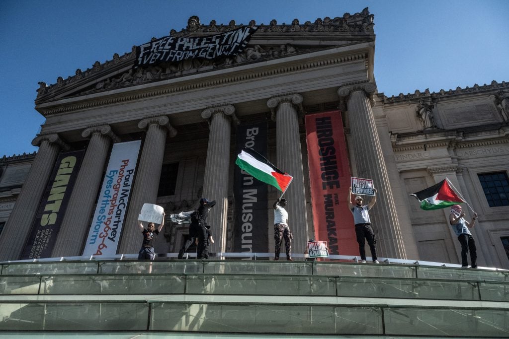 Four protestors, one waving a Palestinian flag, standing on the glass roof at the Brooklyn Museum in New York