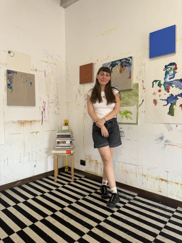 Portrait of artist Cindy Phenix standing in her residency studio with partially finished works on the walls behind her and a black and white tiled floor.