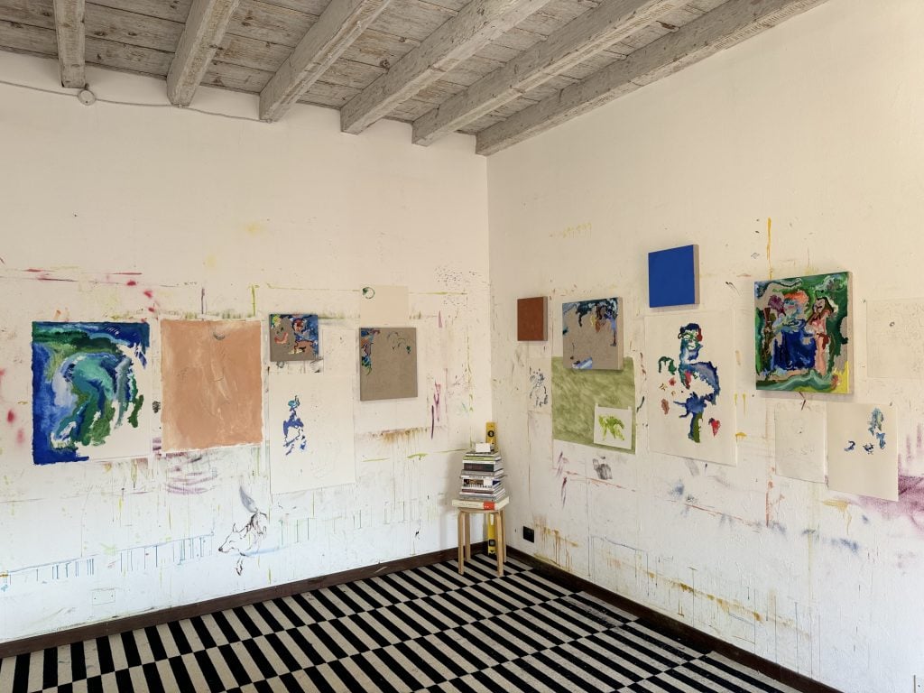 Inside the studio of Cindy Phenix at the Palazzo Monti, with black and white tiled floors and whitewashed beamed ceiling and partially finished works tacked up on the two walls and a stool in the corner with books piled on top.