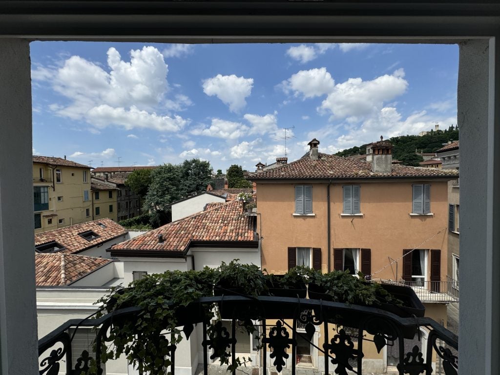 View from the studio of Cindy Phenix at the Palazzo Monti, Brescia, Italy, with a small balcony with planters and a blue sky with clouds and overlooking various stucco buildings with terracotta tiled roofs.