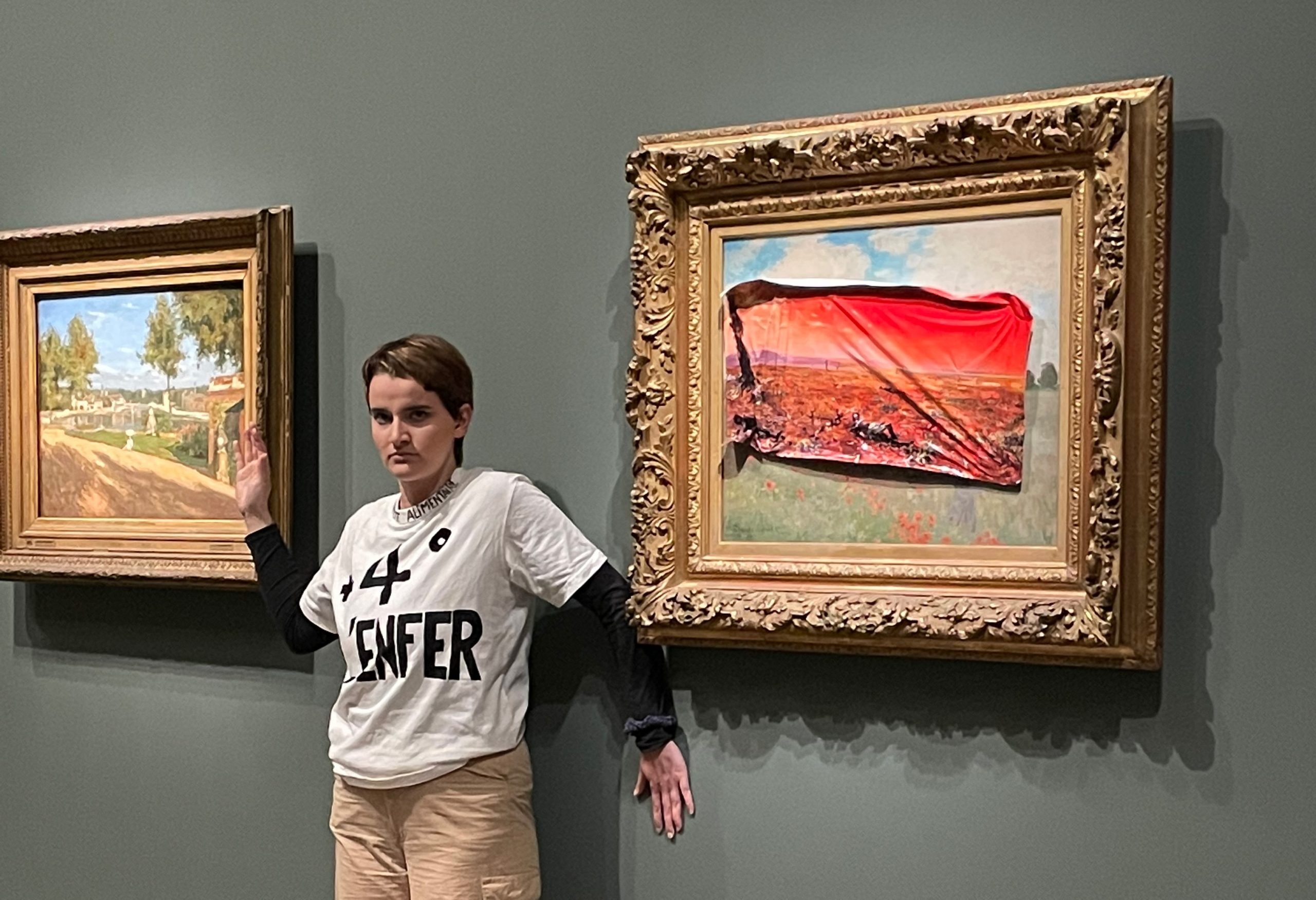 a young woman in a white t-shirt with "4 L'ENFER" written in black pen across the front stands with her hand glued to the wall below a gold framed landscape apinting with a poster scrappily glued over the top showing a more dystopian red-tinged scene of an imagined post-climate collapse environment