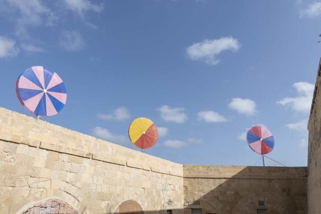 Conrad Shawcross, Beacons (2021). Courtesy of Malta International Contemporary Art Space (MICAS). Three round pinwheel shaped sculptures in multicolor installed across a medieval fortress wall.