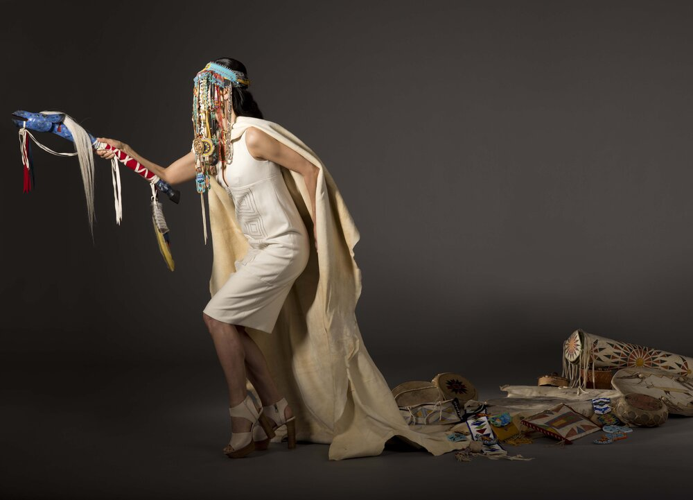 A woman in a grey photo studio space wearing an off-white shift dress and nude heels wearing a traditional Indigenous headdress and holding a ceremonial object with a trail of other cultural objects behind her, part of the must see summer shows list for Artnet.