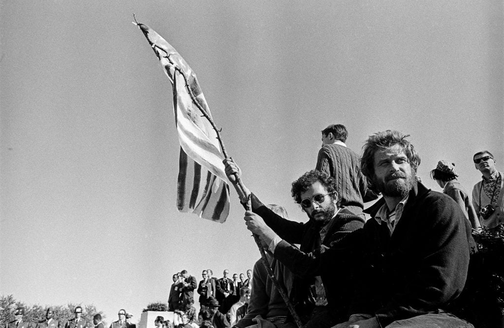 Two men sitting in the foreground of a demonstration, one of them waving an American flag