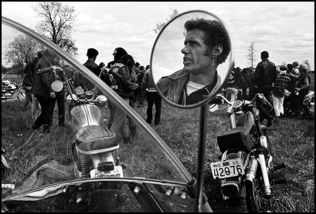 A black and white photo of a man's face reflected in the small rearview mirror of a motorcycle