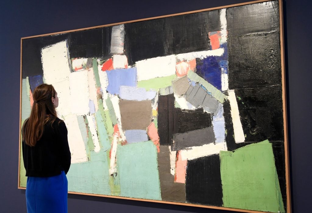 A woman stands in front of artwork featuring broad, textured brushstrokes in a palette dominated by deep blues, muted whites, and earthy browns. The composition is characterized by its geometric abstraction, with overlapping rectangles and forms suggesting a fragmented, aerial view of a stadium and surrounding landscape. 