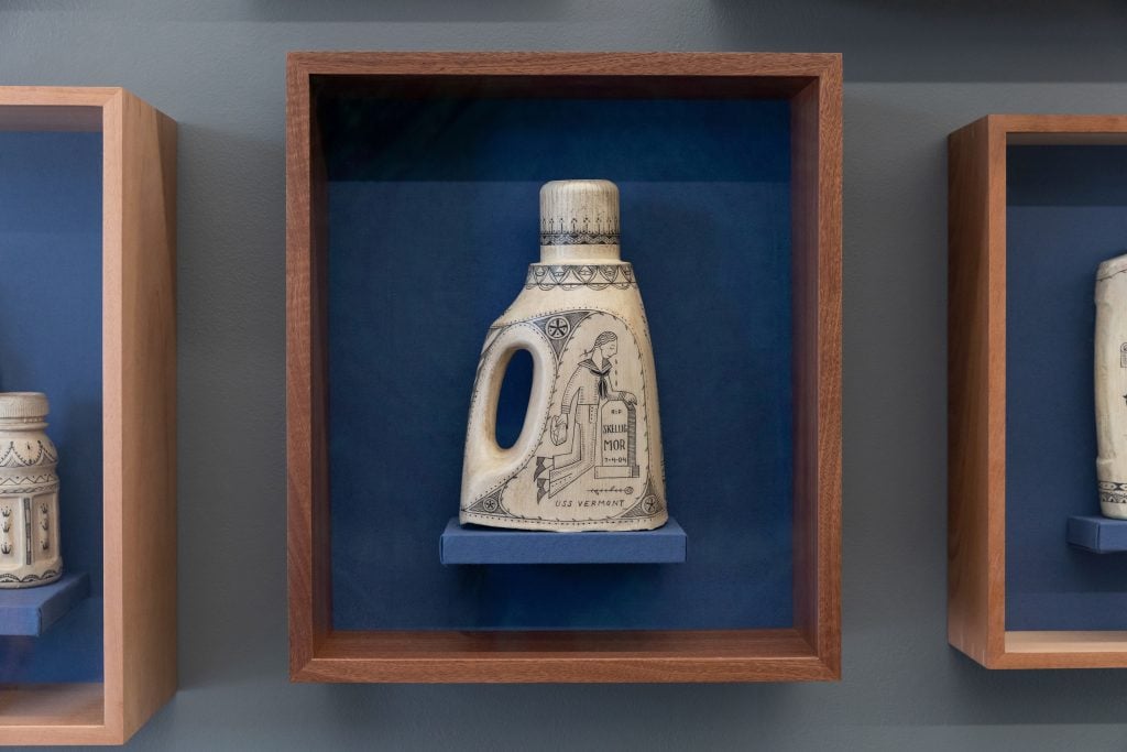 A scrimshaw bottle by Duke Riley depicting a naval officer weeping at a tombstone, on display in a gallery