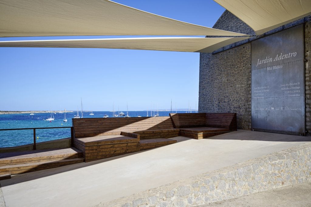 Exterior view of Fundación La Nave Salina in Ibiza, with a view of the Mediterranean dotted with dozens of sailboats in the background, and several cloth triangle awnings stretching out from a stone building that houses the exhibition space.