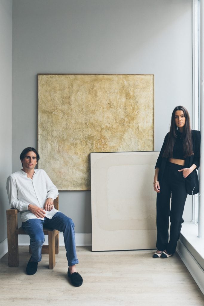 Portrait of Cadogan Gallery Managing Director Freddie Burness seated on the left wearing white button down and blue jeans and Associate Director Katie Burness leaning on a window frame on the right in a black suit standing in front of one abstract pale ochre abstract painting installed on a pale blue wall and a monochrome cream painting leaning on it in front.