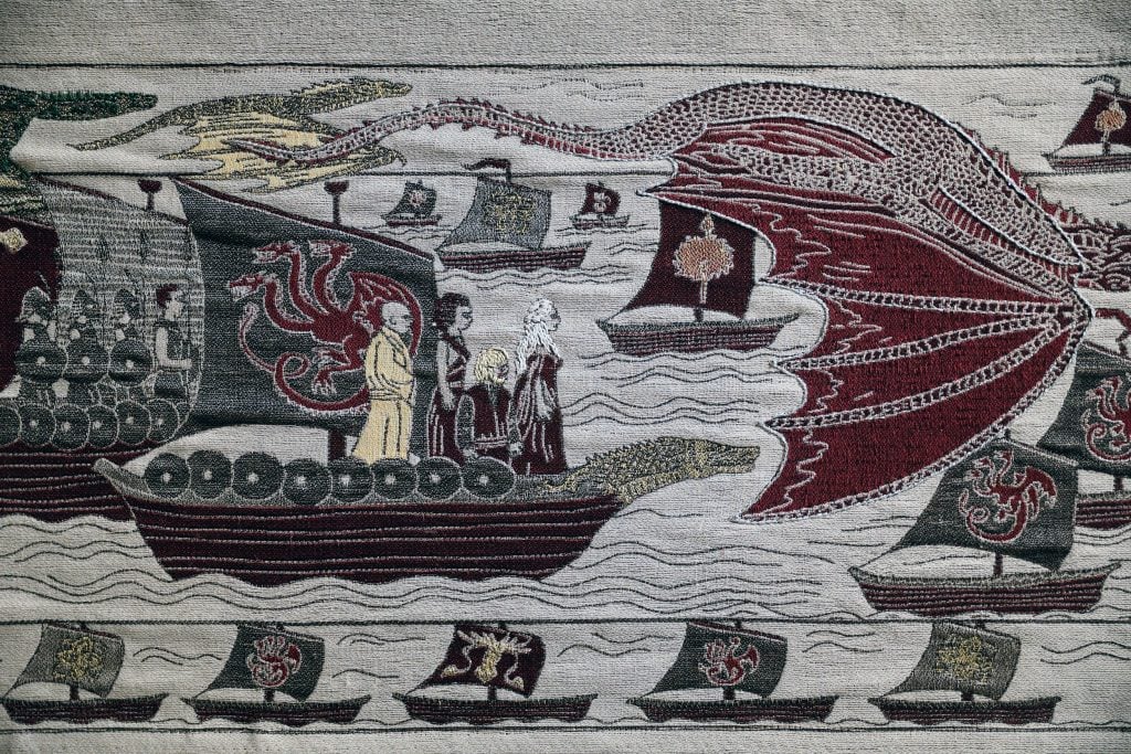 Embroidered tapestry depicting a ship sailing with four people at the helm and a flag with a many-headed dragon, surrounded by other ships