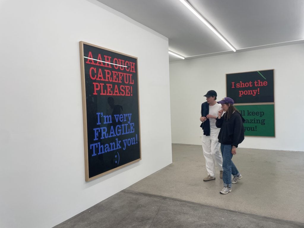 two people walk through an art gallery. the walls are white and there are two large text-based paintings on the walls