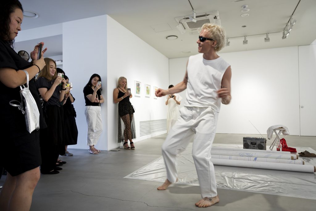 A performer dressed in all white, including sunglasses, is energetically dancing in an art gallery. The audience, mainly dressed in black, watches attentively and records the performance on their phones. The gallery setting includes minimalist white walls with artwork displayed, and a few items, such as rolled-up papers and a boombox, are visible on the floor. The atmosphere is lively and engaging, highlighting the contrast between the performer's dynamic movement and the spectators' focused attention.