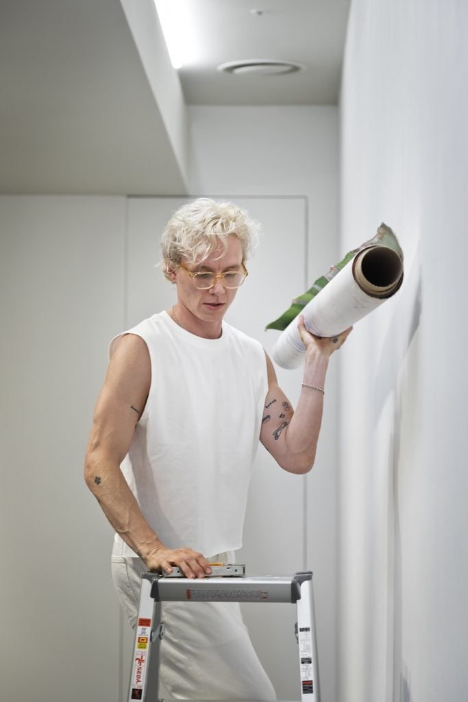 A person with short, curly blonde hair and wearing glasses is standing on a ladder, holding a rolled-up piece of artwork. Dressed in a white sleeveless shirt and white pants, the individual appears focused as they prepare to hang the artwork on a minimalist white wall. The setting is an art gallery, emphasizing the careful and thoughtful process of setting up an exhibition. Various small tattoos are visible on the person's arms, adding a personal touch to their appearance.