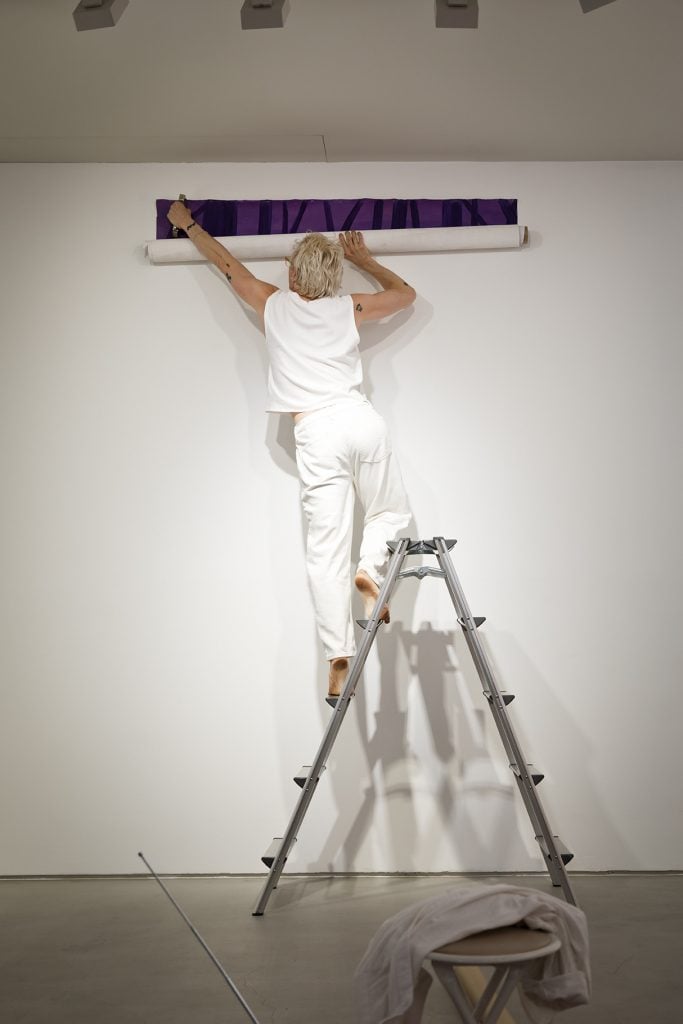 A person with short, curly blonde hair is standing barefoot on a ladder, attaching a rolled-up piece of purple artwork to a white gallery wall. Dressed in a white sleeveless shirt and white pants, they appear focused on carefully positioning the piece. The minimalist gallery setting highlights the preparation process for an exhibition, emphasizing the precision and effort involved. The scene captures the dedication and behind-the-scenes work of an artist or curator setting up for an art show.
