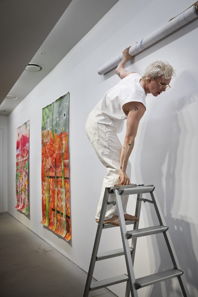A person with short, curly blonde hair, dressed in a white sleeveless shirt and white pants, stands barefoot on a ladder, carefully positioning a rolled-up piece of artwork on a gallery wall. The individual is focused on their task, with colorful, abstract pieces already hanging on the wall behind them. The minimalist gallery setting emphasizes the vibrant artwork, creating a striking visual contrast. The scene captures the careful preparation and dedication involved in setting up an art exhibition, highlighting the artist or curator's meticulous attention to detail.