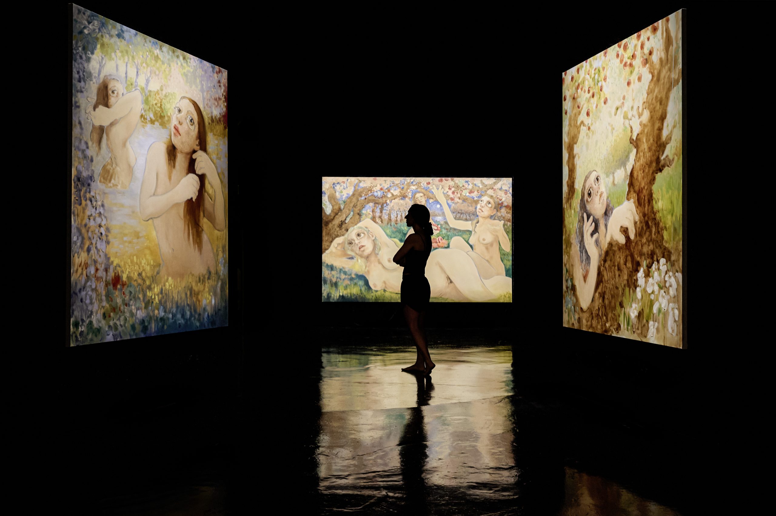 The artist Mai Blanco in shadowed outline at the Fundación La Nave Salinas looking at her large-scale figurative abstract paintings illuminated and reflecting on the black glossy ground.